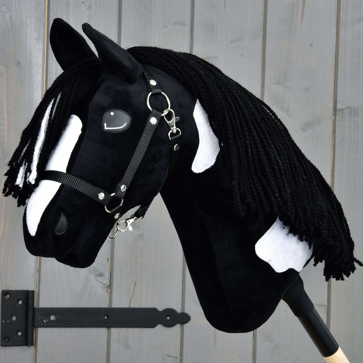 The Hobby Horse LarDen "Hobby Horse Merlin with Black Halter" is a handmade black and white hobby horse with a yarn mane, mounted on a stick and shown against a gray wooden background.