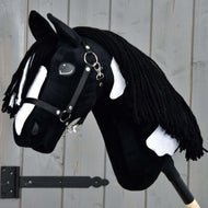 The Hobby Horse LarDen "Hobby Horse Merlin with Black Halter" is a handmade black and white hobby horse with a yarn mane, mounted on a stick and shown against a gray wooden background.