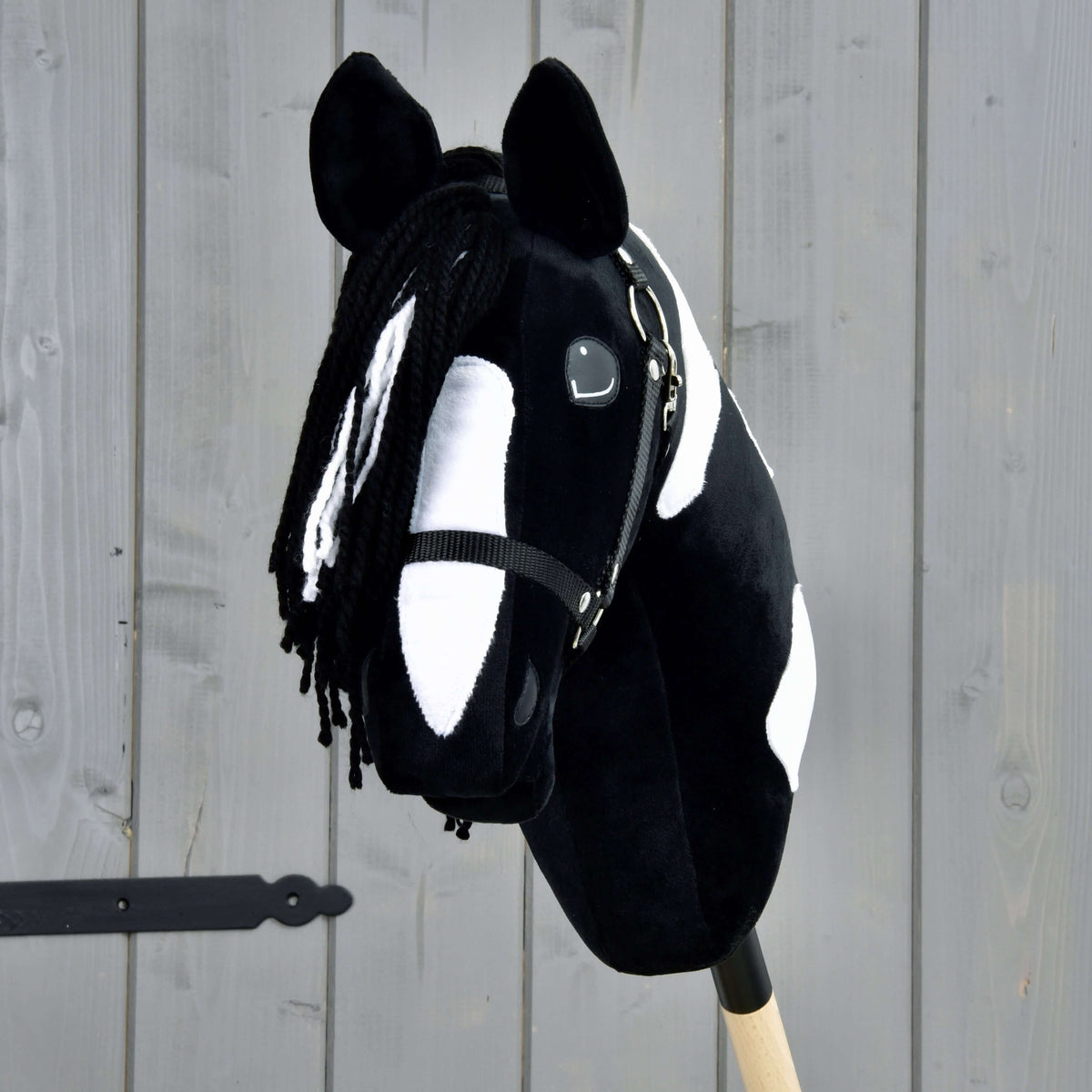The Hobby Horse LarDen "Hobby Horse Merlin with Black Halter" features a plush black and white spotted design with yarn mane, mounted on a wooden stick, and is shown in front of a gray wooden fence, inspired by the Barock Pinto breed.