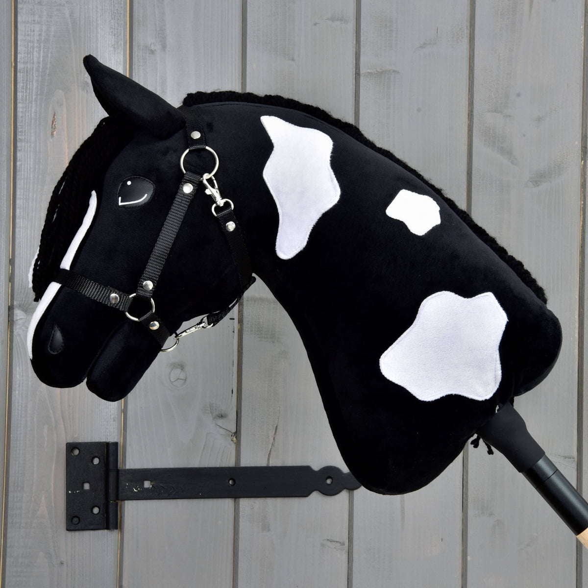 The Hobby Horse LarDen "Hobby Horse Merlin with Black Halter" is mounted on a wooden stick and set against a light gray wooden plank background.