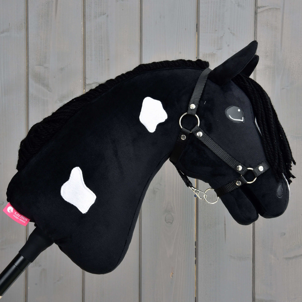 The Hobby Horse Merlin with Black Halter by Hobby Horse LarDen, inspired by the Barock Pinto, features black plush with white patches, a black yarn mane, and bridle, displayed against a wooden plank background.