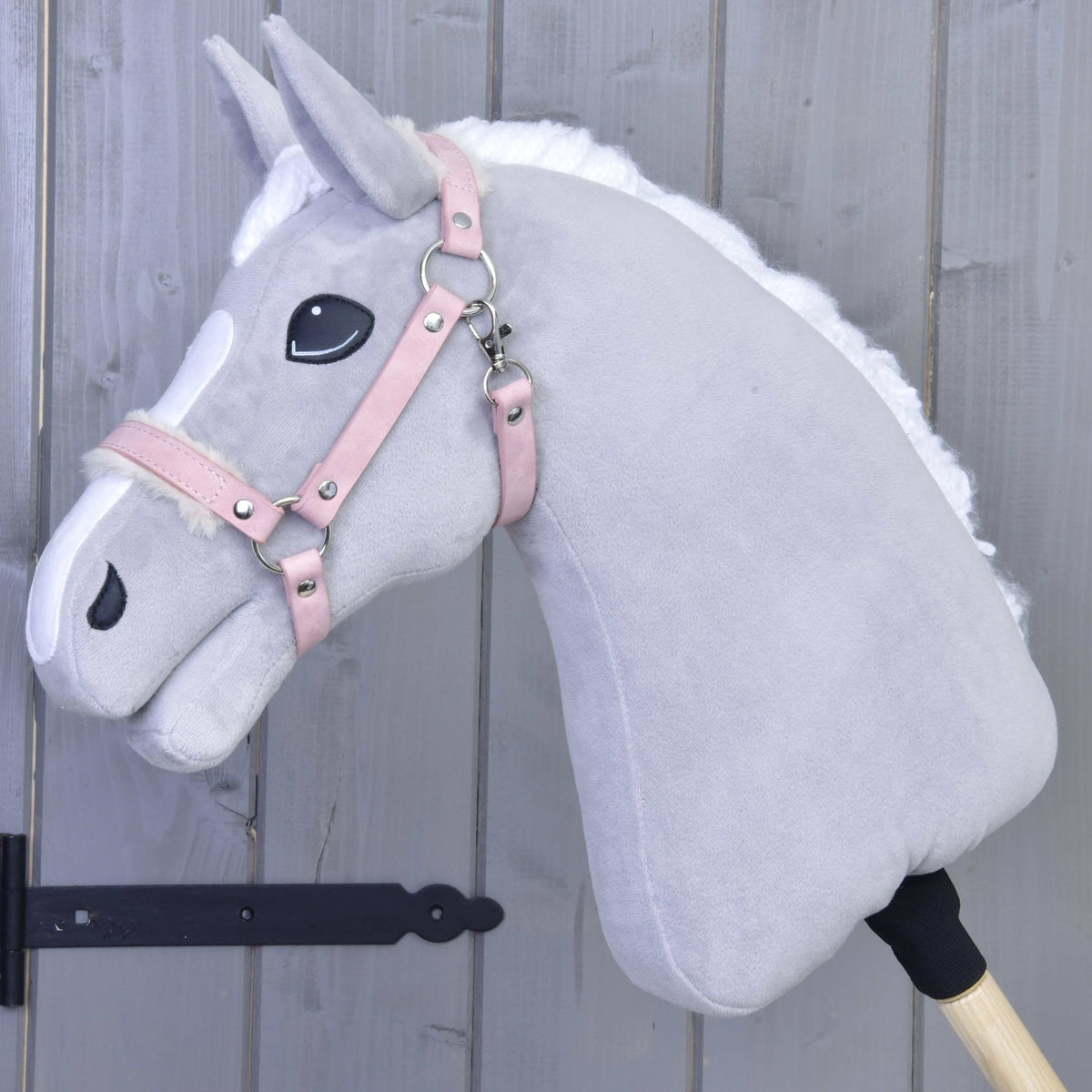 The Hobby Horse LarDen Halter with fur - leatherette Mullbery is a light gray plush hobby horse with a white mane, pink leatherette bridle, and embroidered features mounted on a wooden stick in front of a gray wooden wall.