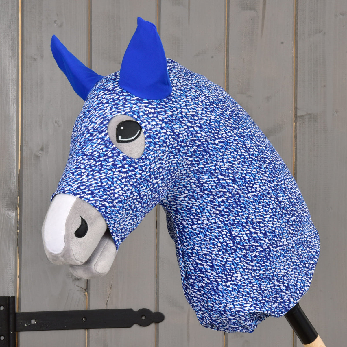 A close-up of the Hobby Horse LarDen "Hobby Horse protective suit See," featuring blue and white PES fabric with blue ears and gray muzzle, mounted on a wooden stick against a gray wooden background.