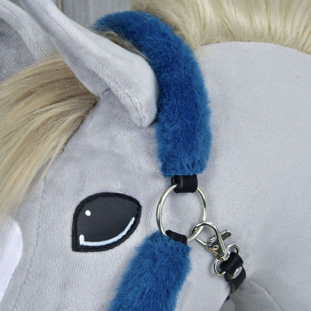 Close-up of a grey plush horse’s head with a cream mane, black and white embroidered eye, and wearing the Hobby Horse LarDen Fur Halter Teddy Petrol in blue with metal rings and clasp.