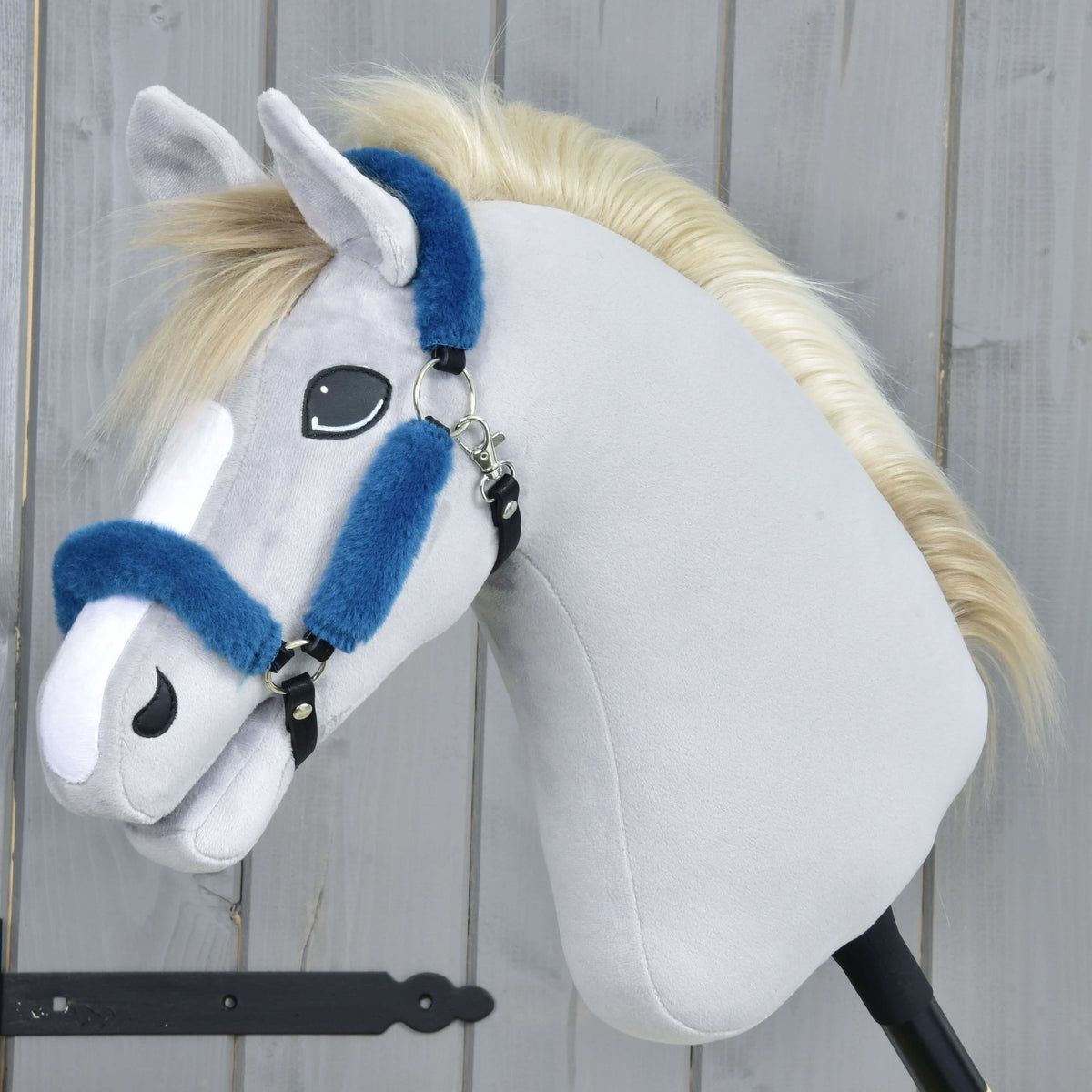 The Hobby Horse LarDen Fur Halter Teddy Petrol is a plush hobby horse featuring light gray fabric, a white muzzle, a blond mane, and a blue leatherette halter on a black stick, displayed before a wooden background.