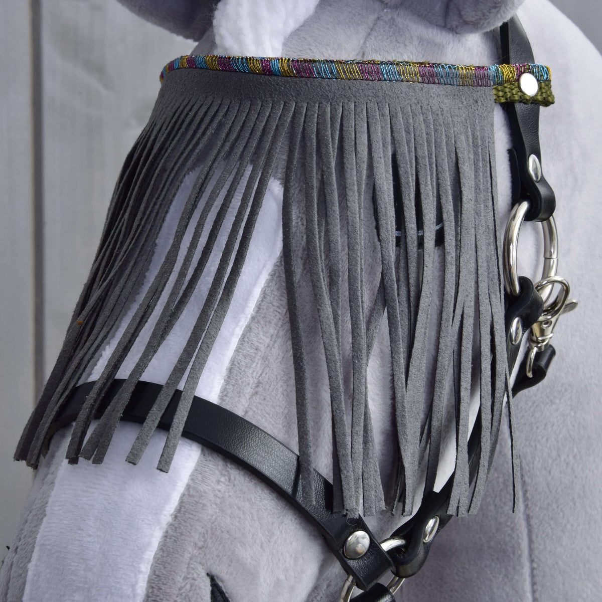 Close-up of a light grey and white horse wearing the Hobby Horse LarDen Halter black with grey fringes, featuring long vertical fringe over its forehead and a colorful trim at the top.