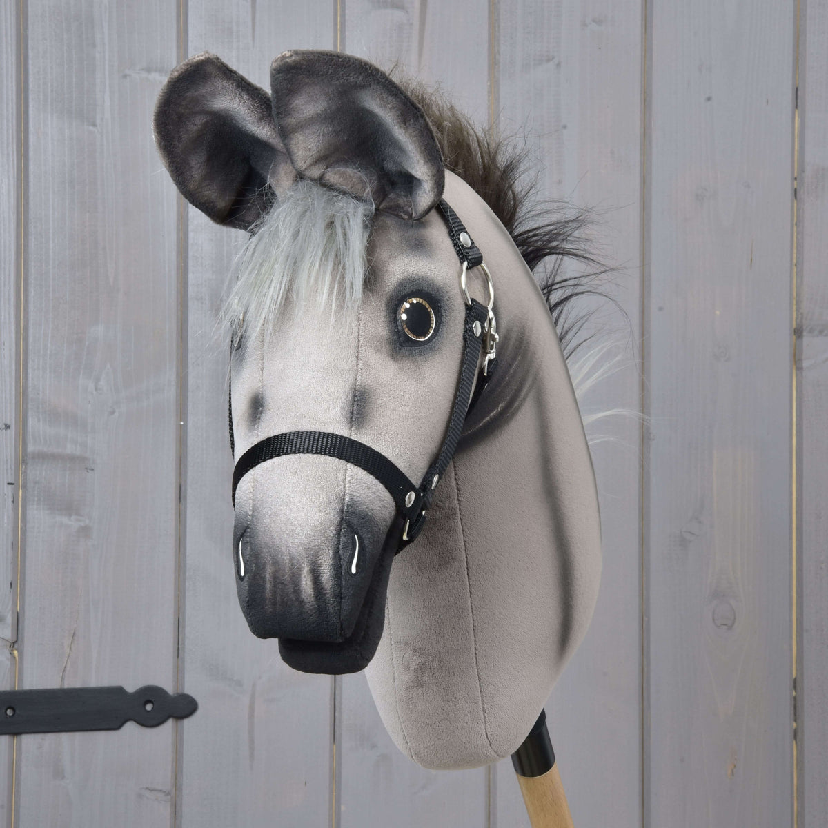 The Hobby Horse Donkey Olin with Black Halter by Hobby Horse LarDen is a gray donkey toy mounted on a wooden stick, featuring painted details and a black harness, set against a light gray wooden panel background.