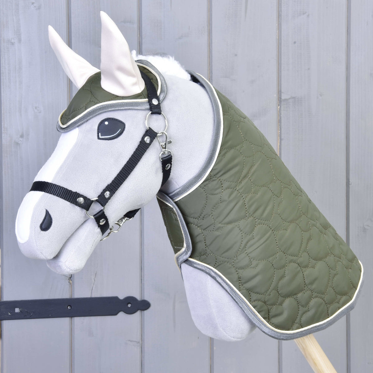 The Set Olivetti by Hobby Horse LarDen is a gray hobby horse with a green quilted blanket and bridle, black halter, white ears, and stylish accessories, shown against a light gray wooden wall.