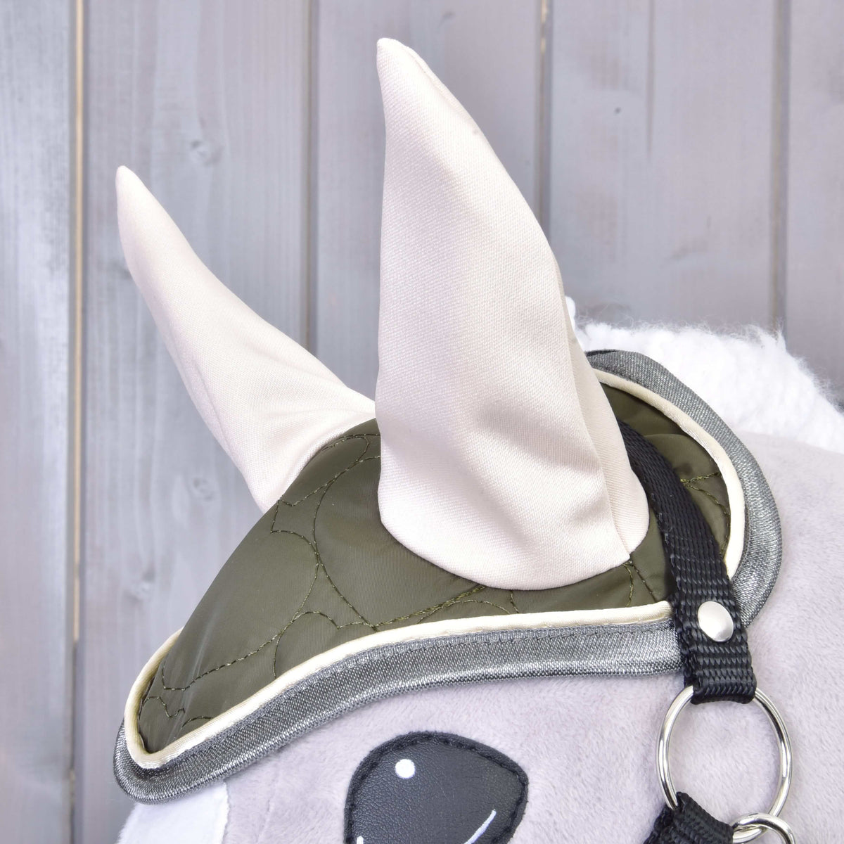 Close-up of a plush toy horse wearing the Set Olivetti by Hobby Horse LarDen: a green quilted ear bonnet with two large light-colored ears, black strap, and metal ring—unique horse accessories on a wooden background.