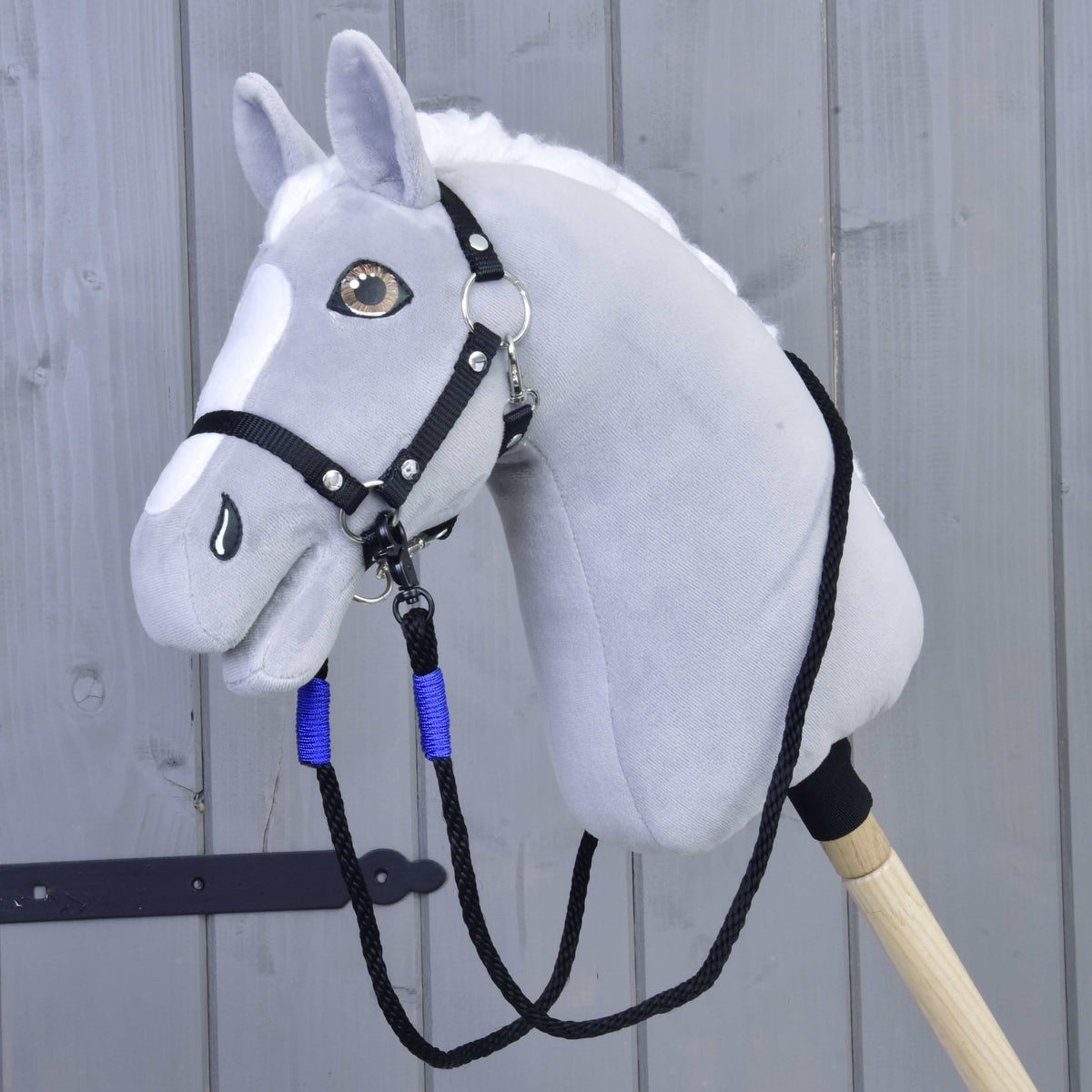 The Hobby Horse LarDen Reins Basic Blue is a gray plush hobby horse head with a black bridle and blue polypropylene rope reins on a wooden stick, shown against a light gray wooden wall background.
