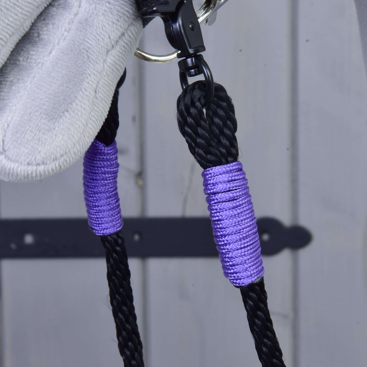 Close-up of Hobby Horse LarDen Reins Basic Lila: black polypropylene handmade reins with purple accents and a silver clip, set against a light gray wooden surface with a black metal hinge.