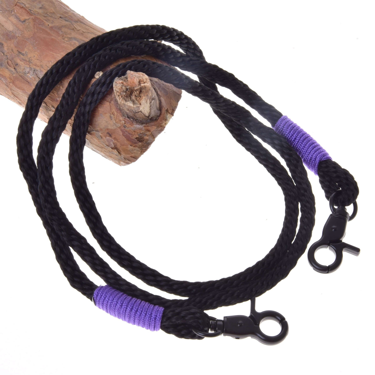 The Reins Basic Lila by Hobby Horse LarDen features a black braided polypropylene rope with purple-wrapped ends and black swivel clips, perfect as handmade or hobby horse reins, shown coiled elegantly on a wooden log.