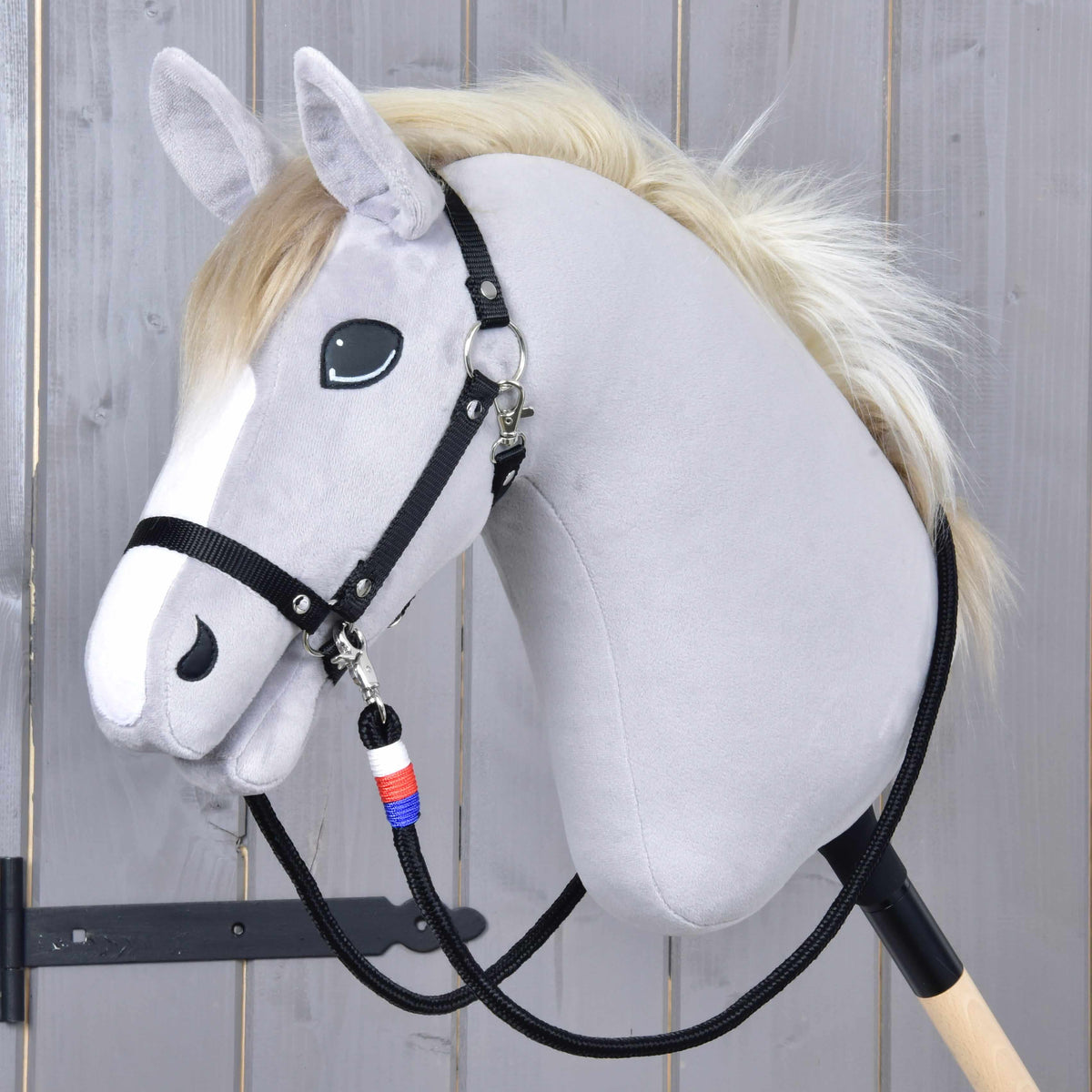 The Hobby Horse LarDen plush hobby horse features a light gray face, black bridle, white mane, and is mounted on a wooden stick. It comes with handmade "Reins Emblem CZ" adorned with a blue, white, and red charm.