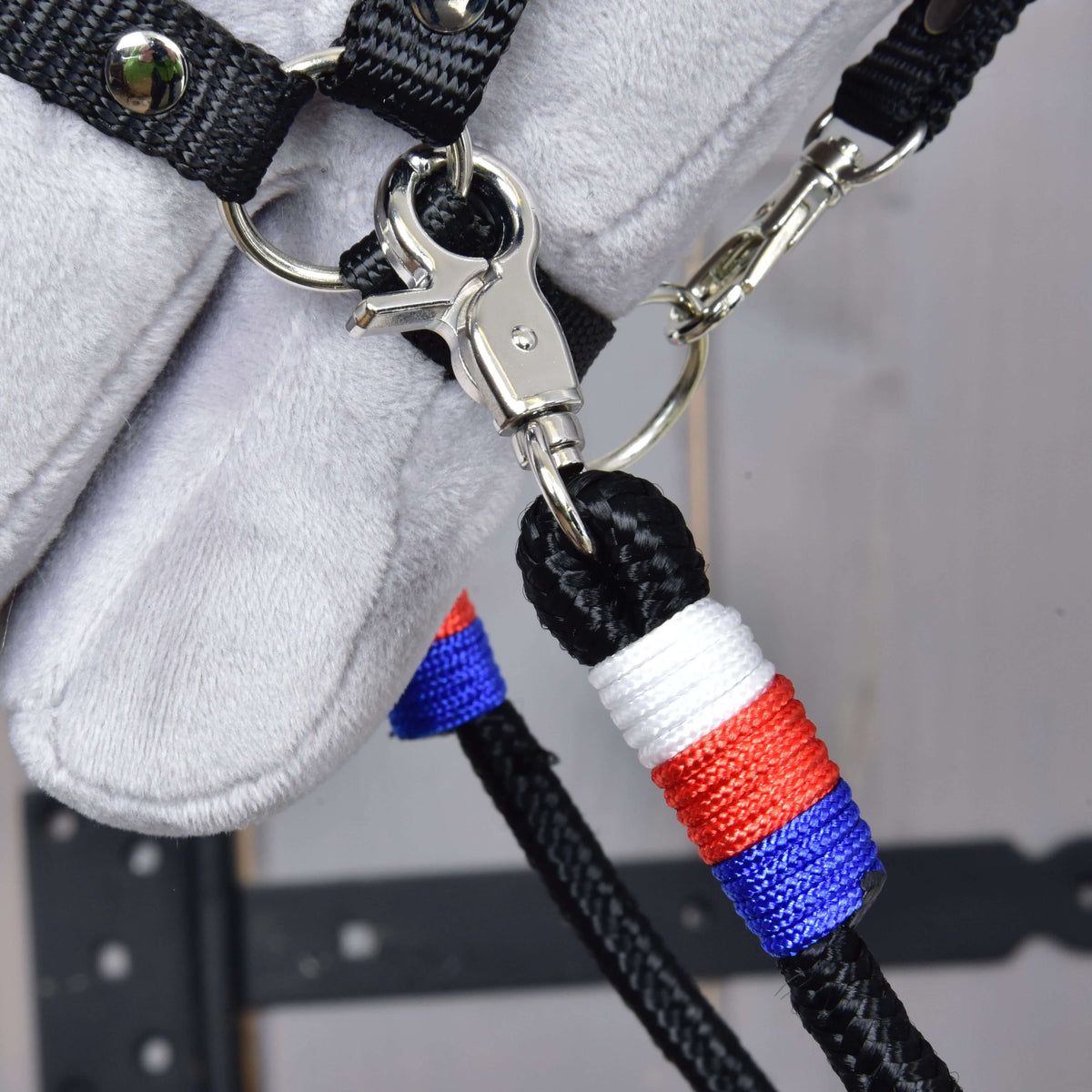 Close-up of the Reins Emblem CZ by Hobby Horse LarDen: handmade black polypropylene rope reins with a metal clasp and red, white, and blue thread wrapping. The horse’s gray fur appears in the background.