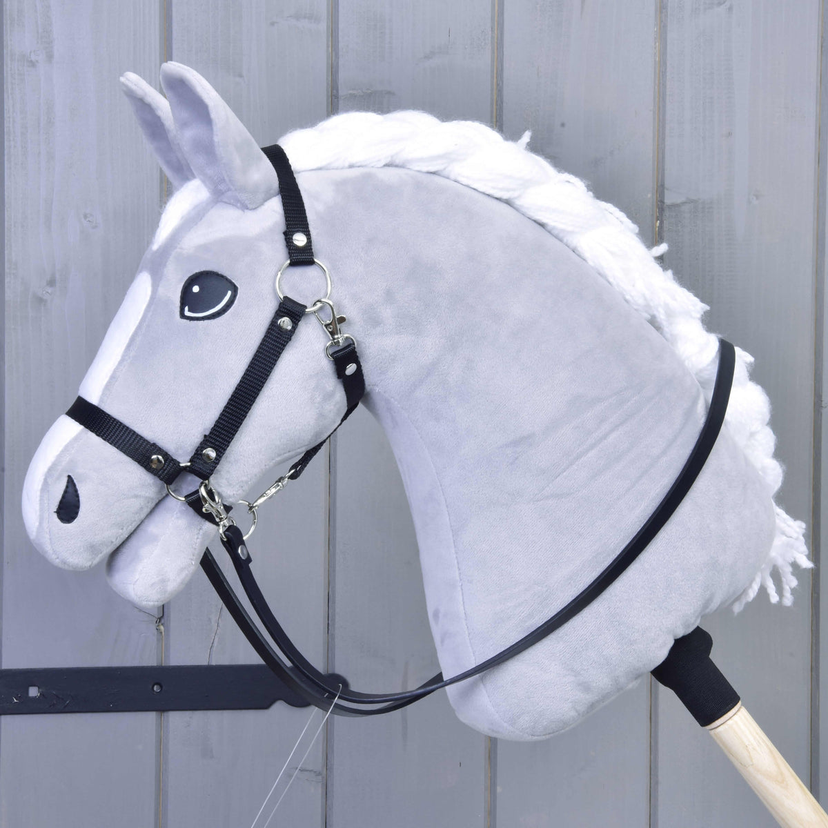 The Hobby Horse LarDen Reins Black leatherette is a gray plush hobby horse with a white mane, bitless bridle, and quality black leatherette reins on a wooden stick, shown against a gray wooden panel background.