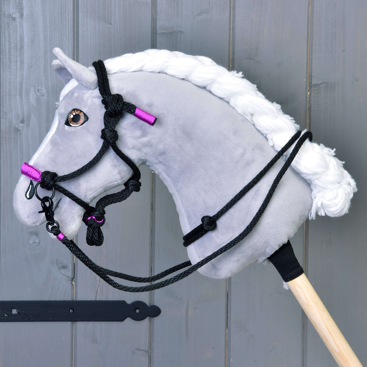 The Hobby Horse LarDen Rope Halter Violette is a gray hobby horse with a white mane, black bridle and rein details, purple accents, and a stylish rope halter, mounted on a wooden stick against a gray wooden wall.