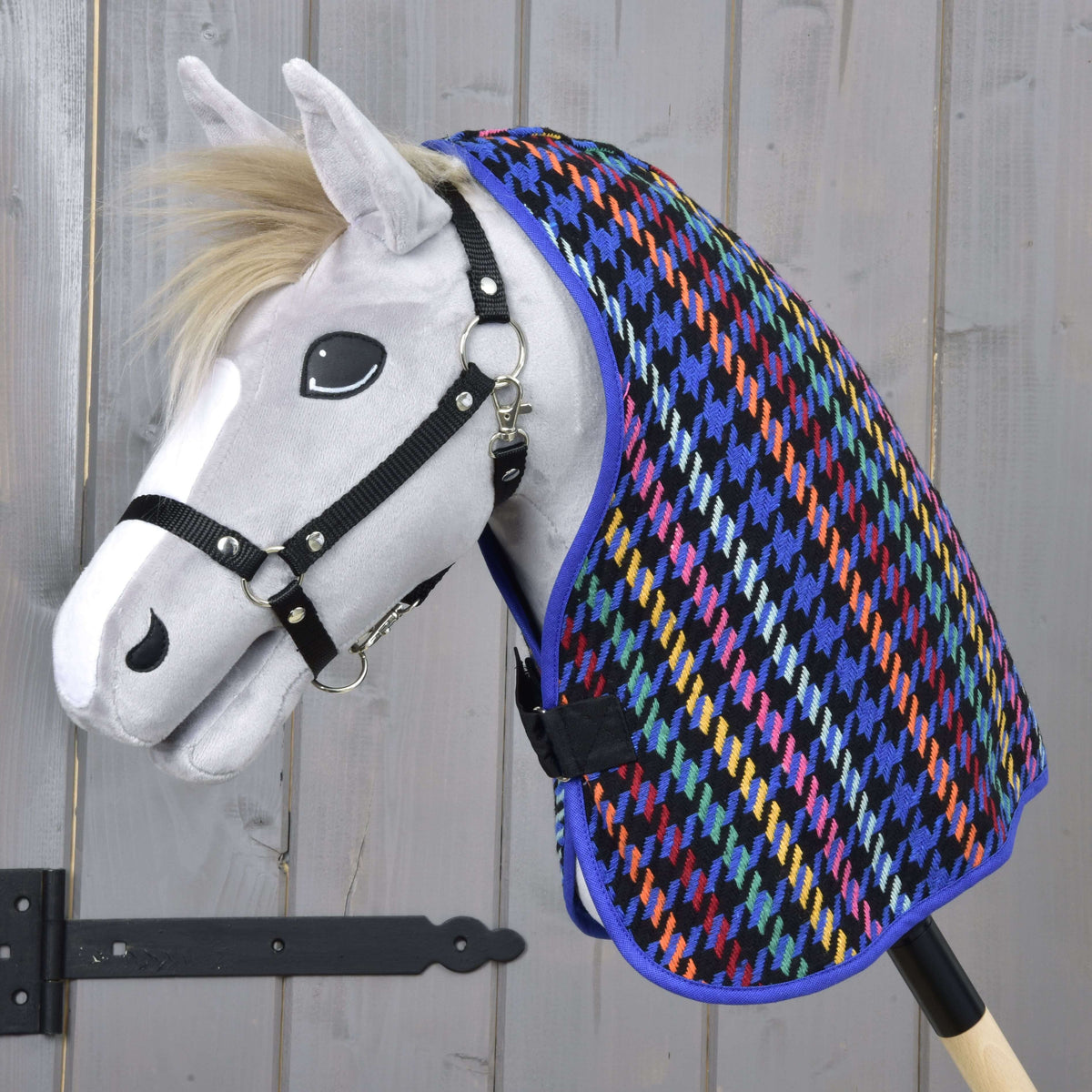 A white toy hobby horse with a black halter wears the colorful Stable Blanket Peru by Hobby Horse LarDen, featuring a houndstooth pattern, set against a light wooden background.