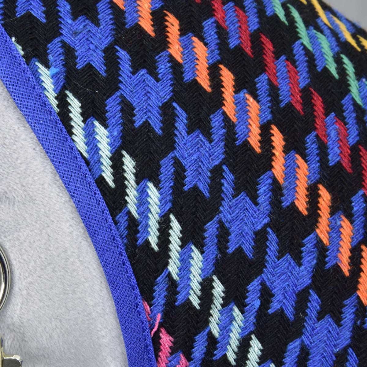 A close-up of the Hobby Horse LarDen Stable Blanket Peru shows a colorful houndstooth pattern in blue, orange, red, yellow, and light green on black, with blue trim and part of a gray background visible.