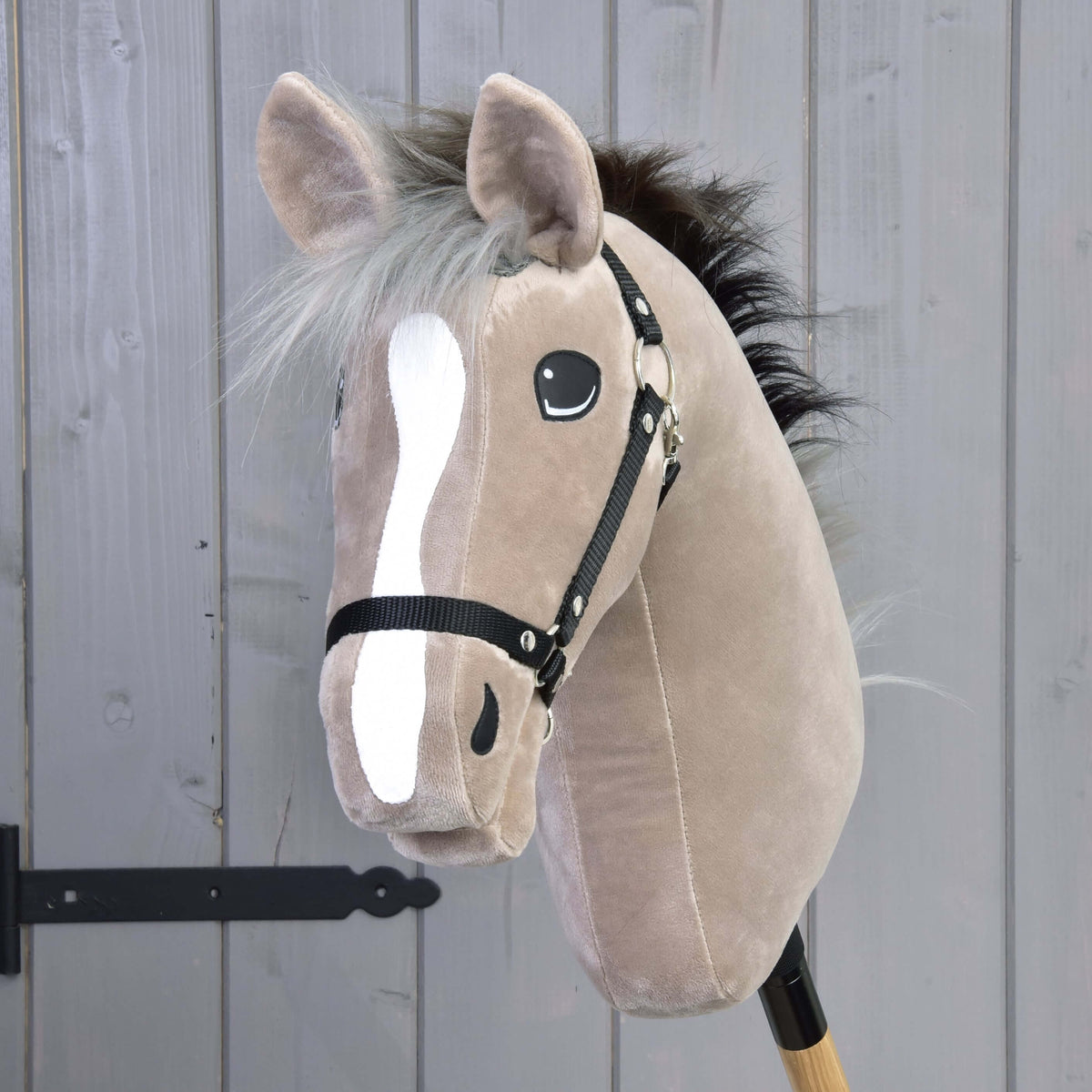 The Hobby Horse LarDen Piccolino with Black Halter is a beige plush hobby horse featuring a white blaze, black button eyes, black mane, and bridle on a wooden stick—perfect for pretend dressage.