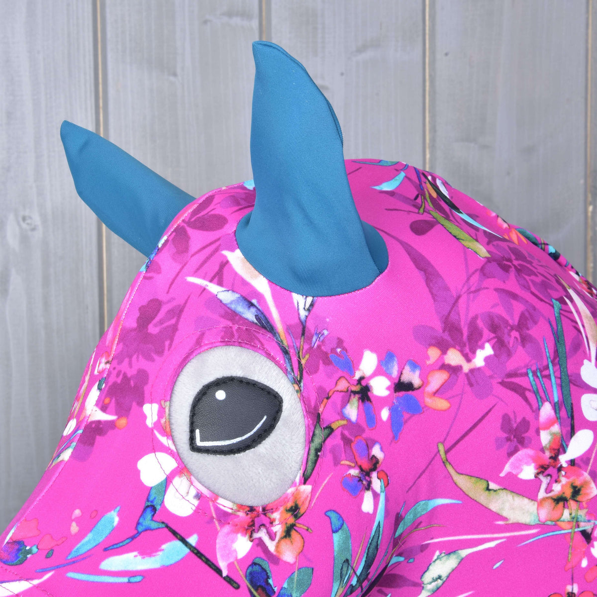The Hobby Horse LarDen "Hobby Horse protective suit Pink Beauty (Lycra)" features pink, floral-patterned fabric, blue Lycra ears/horns, and a large gray and black embroidered eye, shown against a light wooden background.