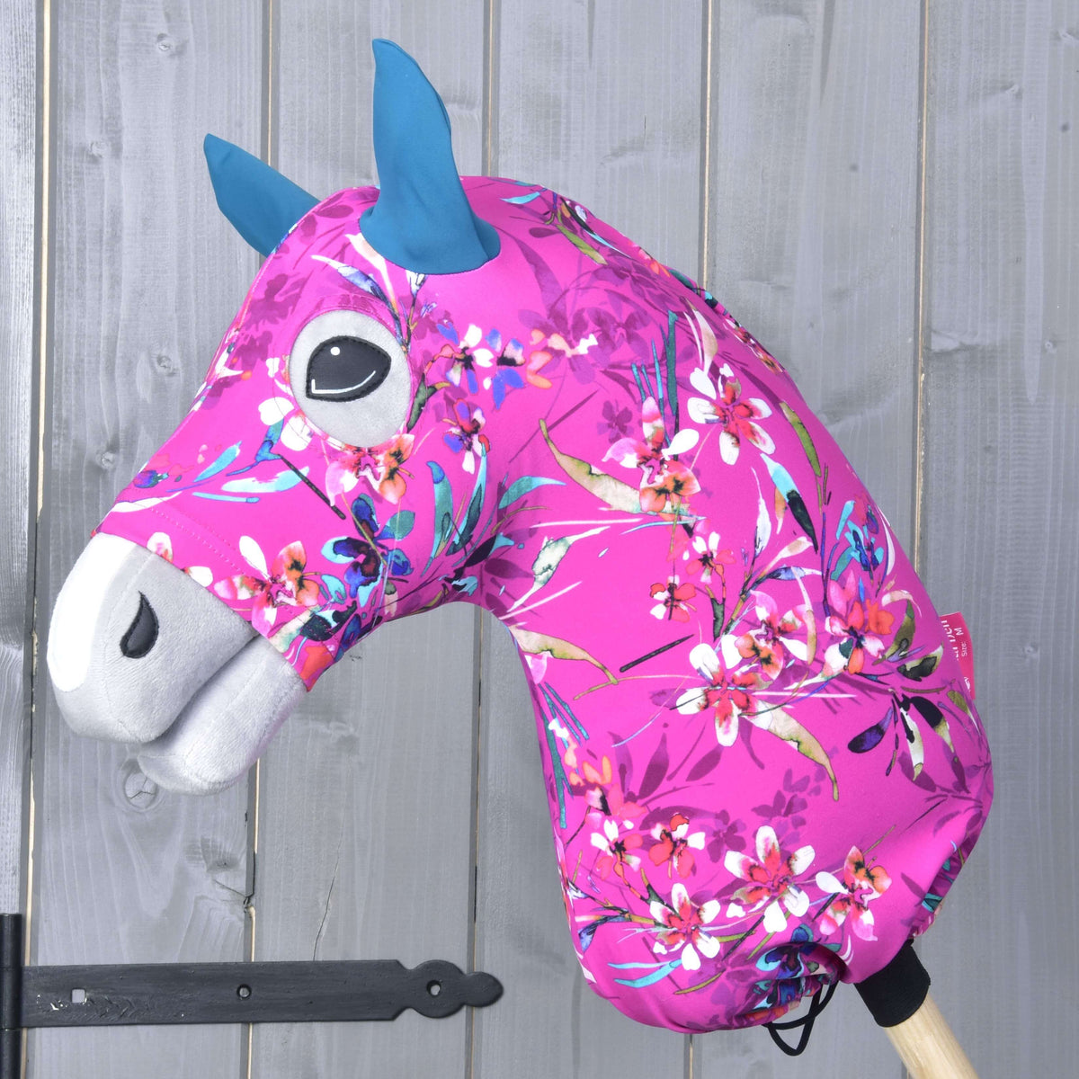 The Hobby Horse LarDen "Hobby Horse protective suit Pink Beauty (Lycra)" features bright pink floral fabric, blue ears, and a white snout, shown mounted on a wooden stick in front of a gray wooden wall.