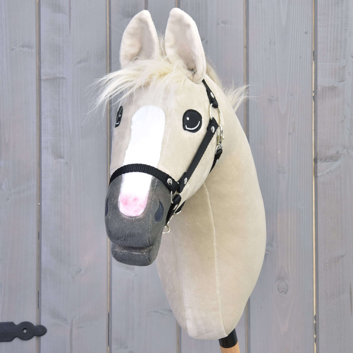 The Hobby Horse LarDen Pixie with Black Halter features a beige coat, white blaze, light mane, and subtle metallic shine, standing before a wooden gray fence.