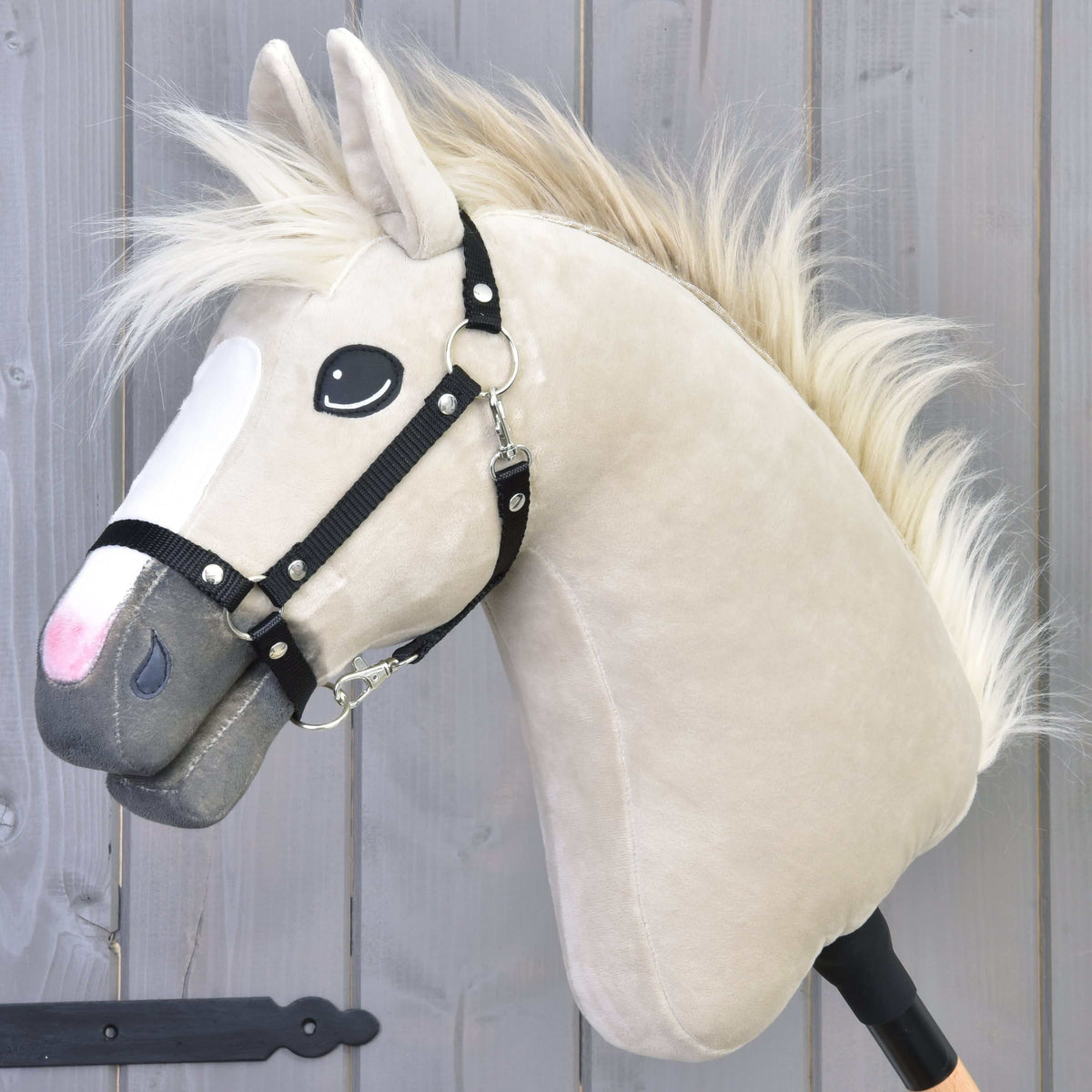 The Hobby Horse LarDen Pixie, featuring a beige coat, white mane, black halter, and expressive eyes on a stick, stands against a gray wooden background, alluding to the Akhal-Teke breed’s legendary metallic gleam.