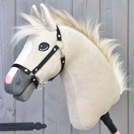 The Hobby Horse LarDen Pixie, featuring a beige coat, white mane, black halter, and expressive eyes on a stick, stands against a gray wooden background, alluding to the Akhal-Teke breed’s legendary metallic gleam.