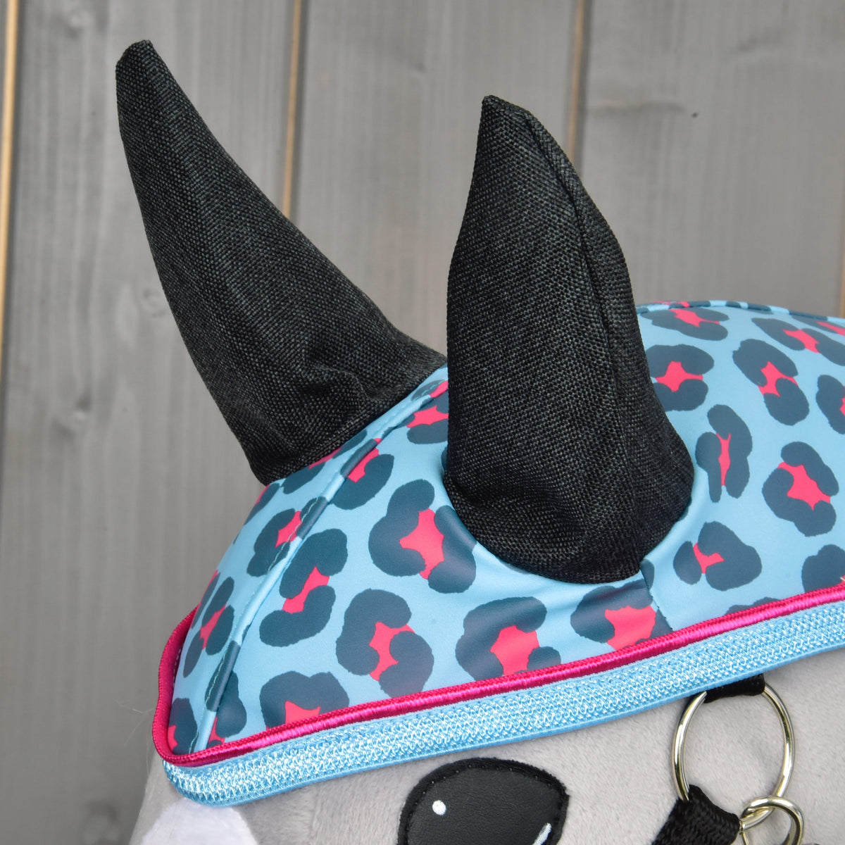 A close-up of the Hobby Horse LarDen "Raincoat Blue Leopard" plush toy in a blue and pink leopard-print hat with black horns, ready for adventure in any weather, set against a wooden background.