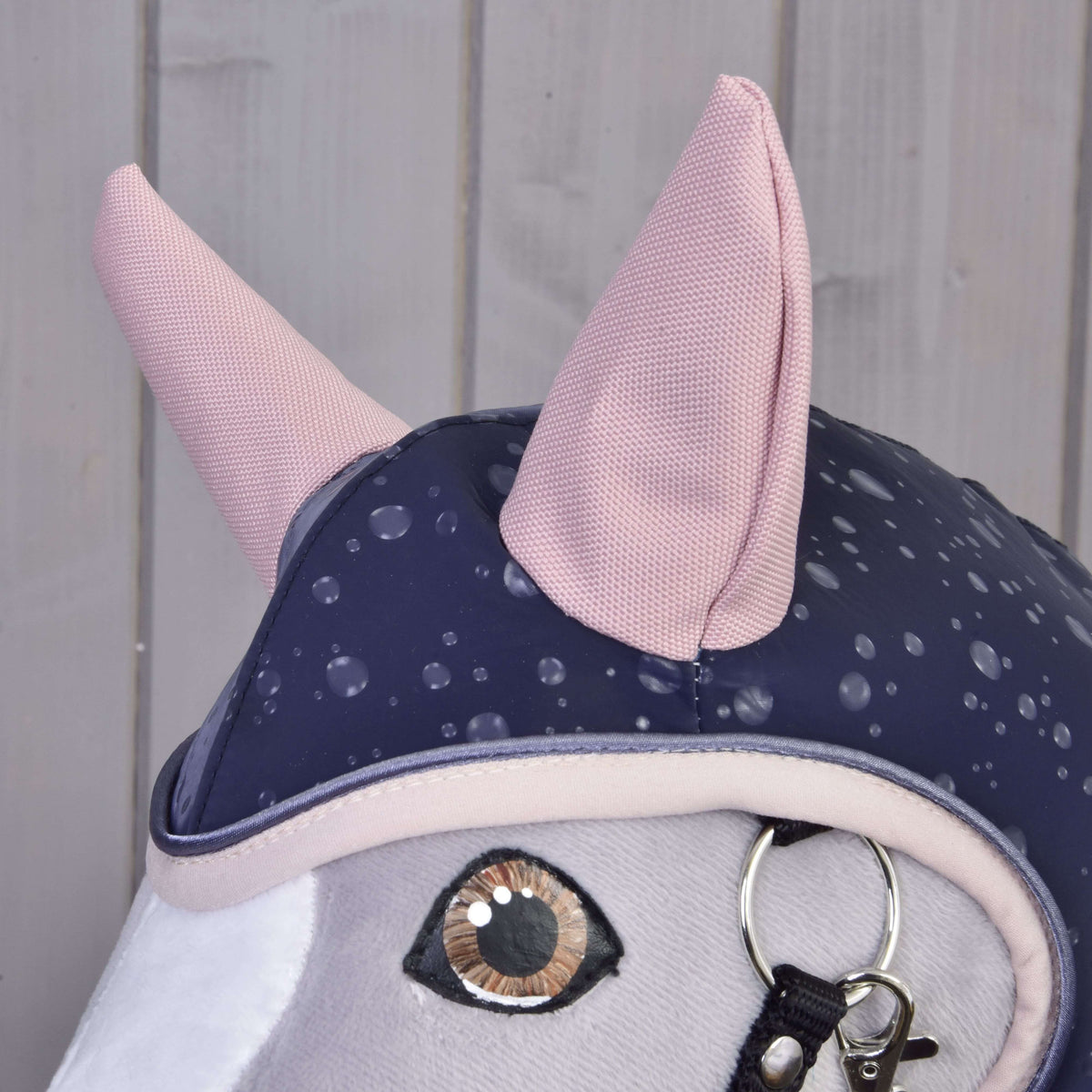 Close-up of Hobby Horse LarDen's Raincoat Pinkie Blue (softshell): plush horse head with large embroidered eye, two pink ears, a blue polka-dot riding cap, and bridle with metal rings. Light wooden panel background.