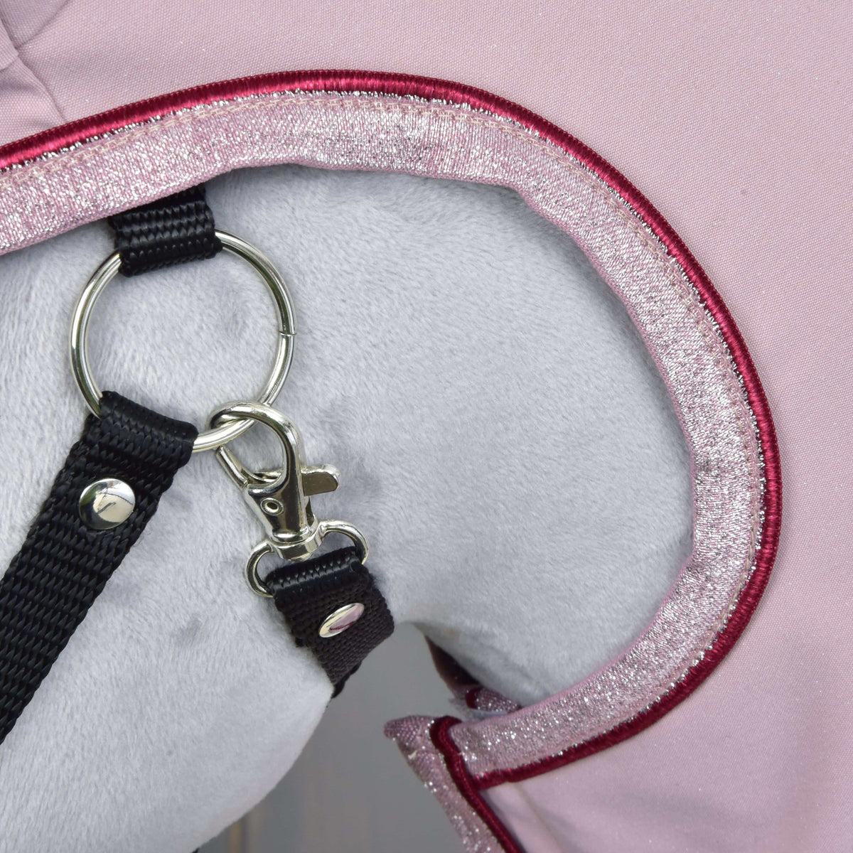 A close-up of a white plush animal wearing the pink, sparkly-edged Raincoat Veloria by Hobby Horse LarDen, with a black leash attached to a silver ring and clasp.