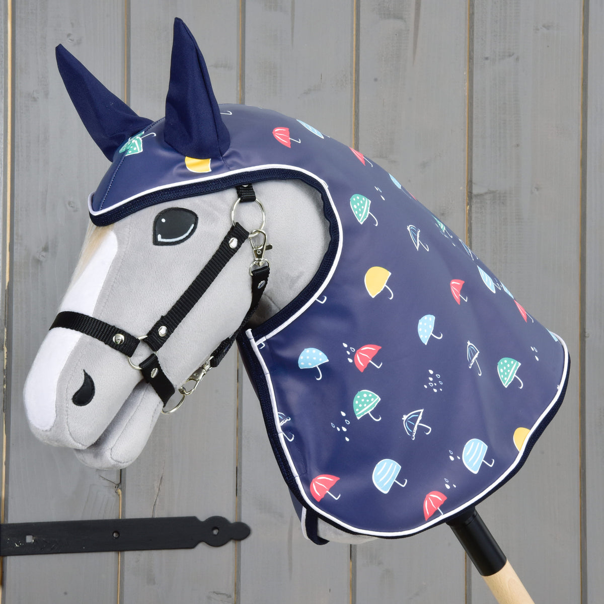 The Hobby Horse LarDen "Raincoat Rainy" is a plush hobby horse with a horse head on a wooden stick, wearing an all-weather blue raincoat hood with colorful umbrella patterns and dark blue ear covers, shown by a gray wooden wall.