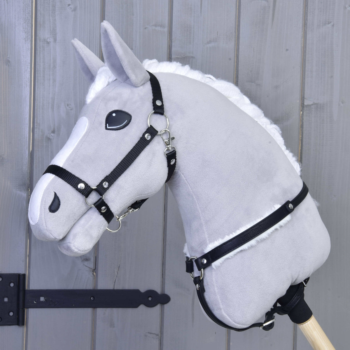 The Hobby Horse LarDen Breastgirth with fur Espresso is a plush gray hobby horse with a white mane, black bridle, and soft fur-lined breastgirth on a wooden stick, shown against a light wood-paneled wall.