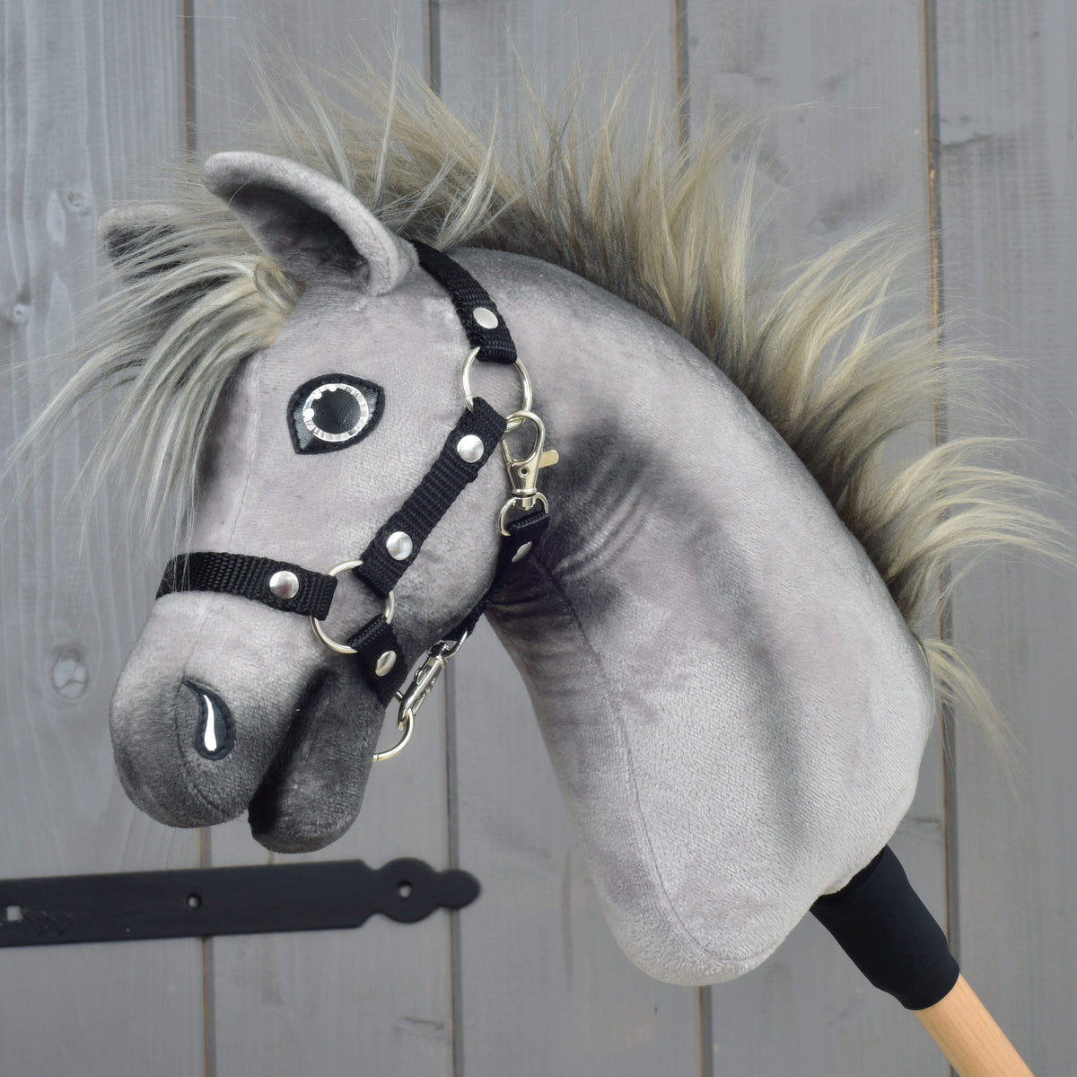 The Hobby Horse LarDen “Hobby Horse Prestige with black halter, size XS” features a gray plush design with a spiky mane and black halter, inspired by the elegance of an English Warmblood. It is mounted on a wooden stick and displayed against a wooden panel background.