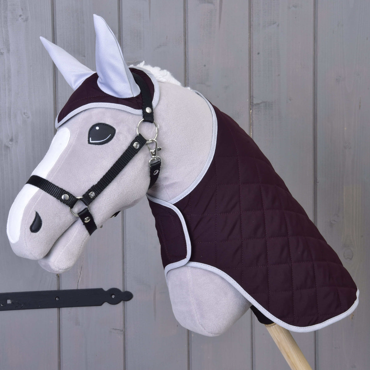 The Hobby Horse LarDen Set Purple features a gray hobby horse toy with a quilted dark purple blanket, bridle, and ear bonnet, all mounted on a wooden stick and shown against a wooden plank background.