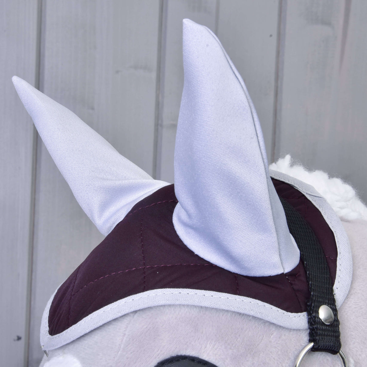 Close-up of Hobby Horse LarDen’s Set Purple: a plush horse head with large gray ears, maroon and gray quilted bridle, and ear bonnet. Vertical light gray wooden panels in the background.
