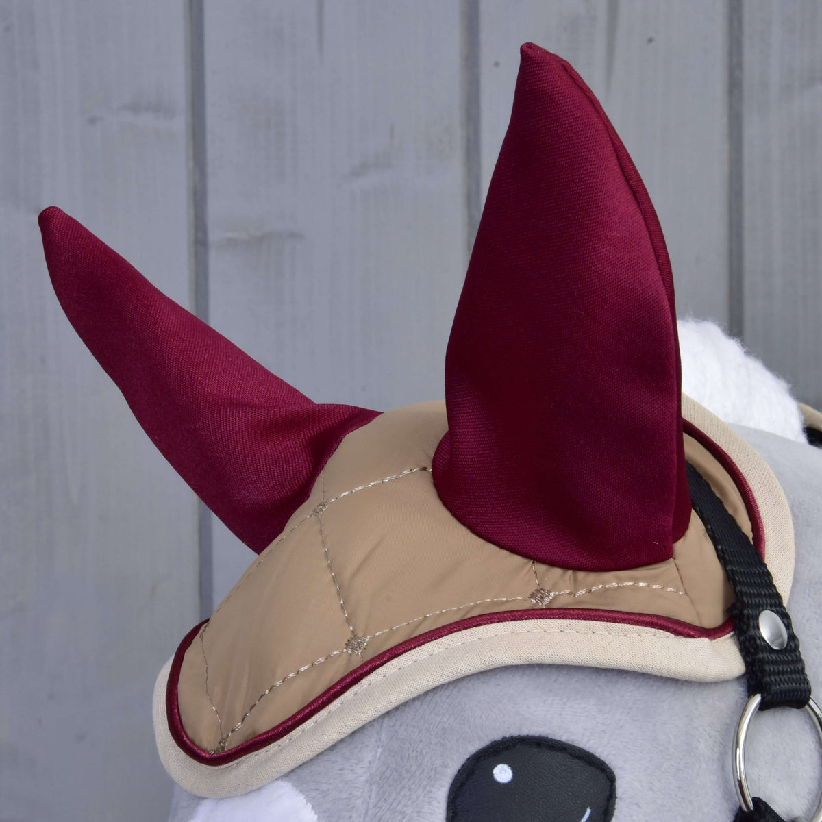 A close-up of the gray Hobby Horse LarDen Set Roma plush toy, featuring a tan bridle and maroon ear bonnet, set against a gray wooden background.