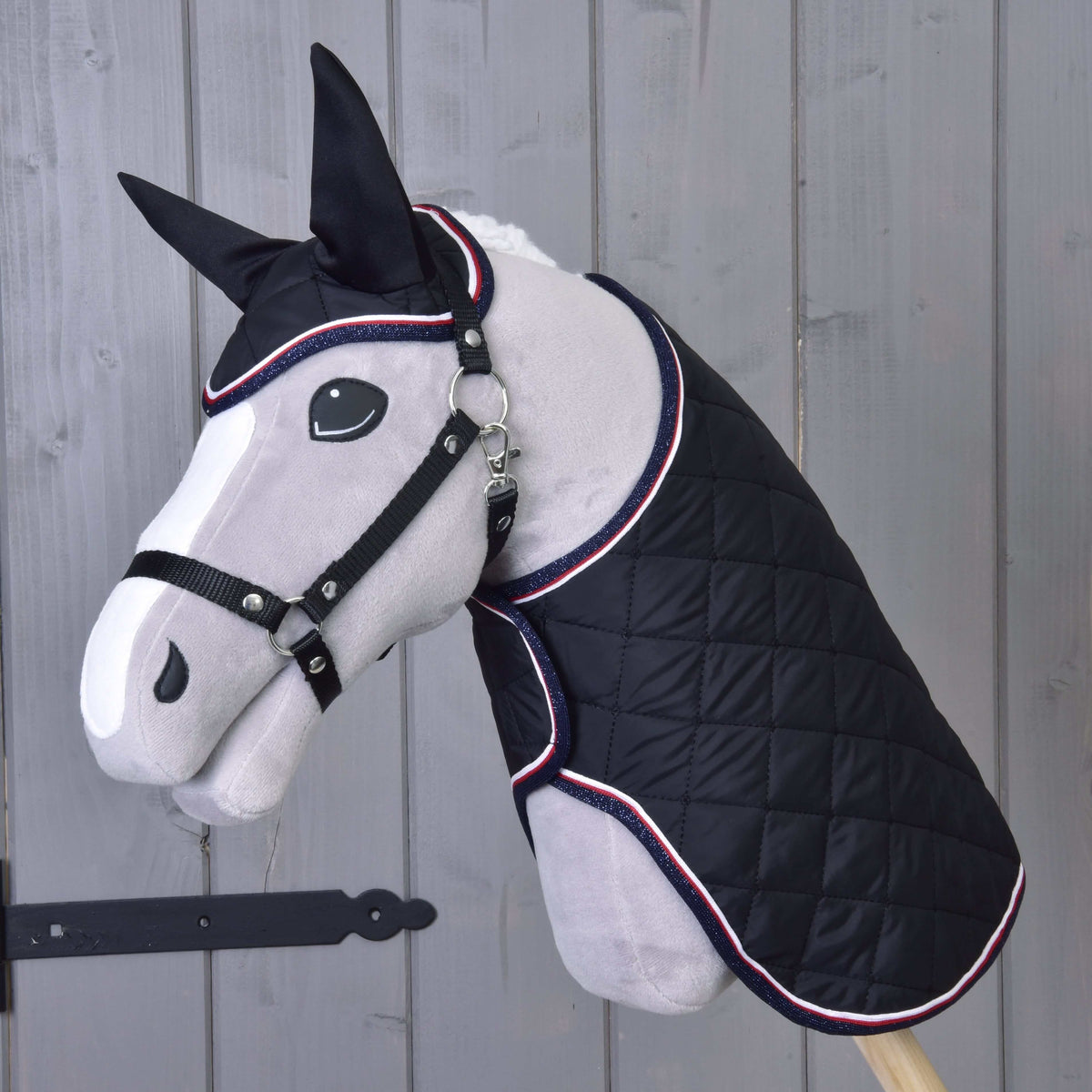 A gray plush horse mannequin models the Hobby Horse LarDen Set Royal, a black quilted blanket and matching ear bonnet. The backdrop is a light wooden wall with a black metal latch on the left.