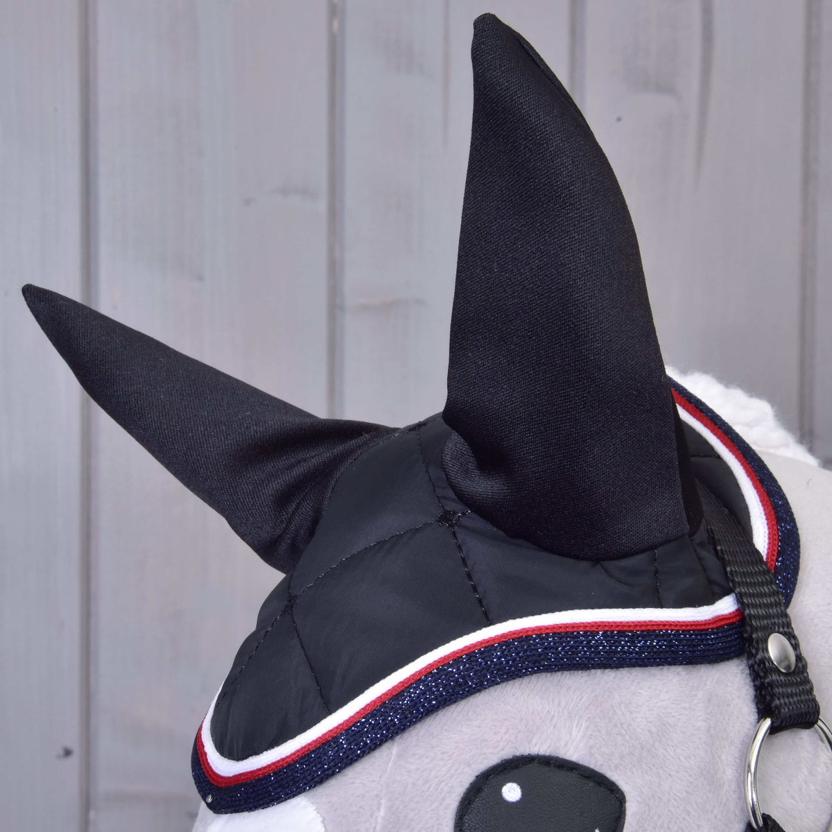 Close-up of Hobby Horse LarDen's Set Royal: a hobby horse head with black fabric ears, a stylish ear bonnet featuring red, white, and blue trim, grey face, and black bridle—an eye-catching accessory on light wood background.