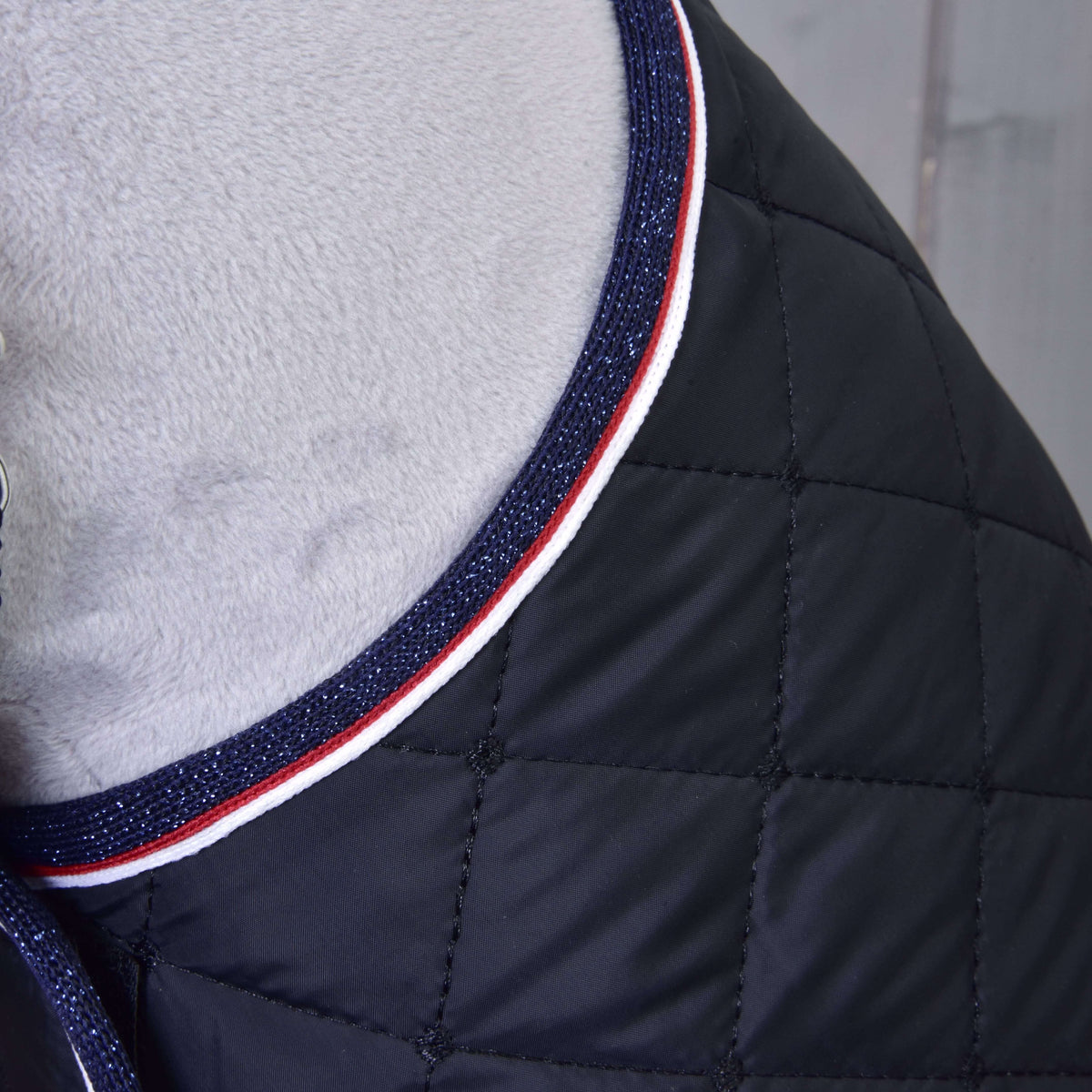 Close-up of the Set Royal quilted black blanket by Hobby Horse LarDen, featuring a diamond pattern and navy blue trim with red and white stripes, elegantly draped over a soft light gray surface—perfect as a horse accessory.