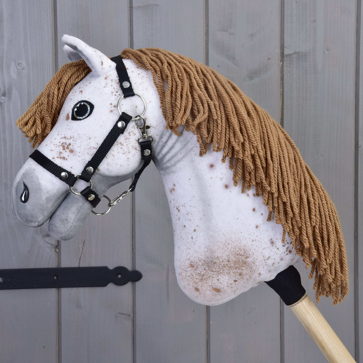 A close-up of Hobby Horse LarDen’s Hobby Horse Rufus with a white face, brown yarn mane, black halter, and speckled brown spots, set against a wooden panel background.