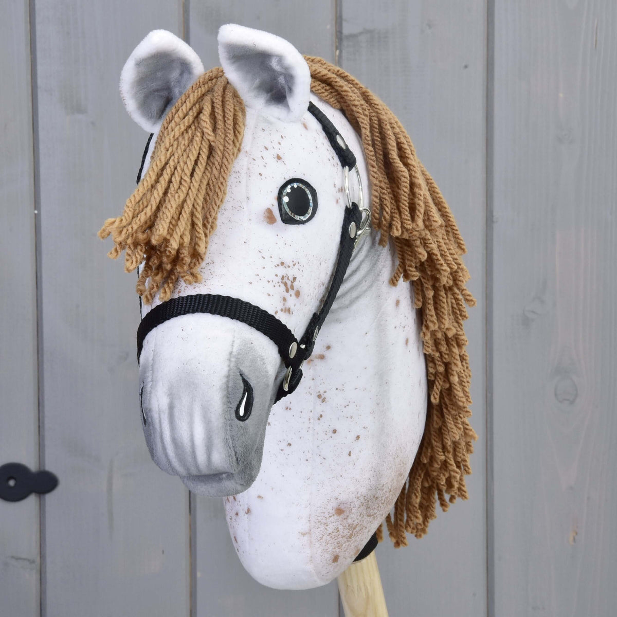 The Hobby Horse Rufus with Black Halter by Hobby Horse LarDen features a handmade white plush face, brown yarn mane, black halter, painted details, and is mounted on a wooden stick against a gray wood panel background.