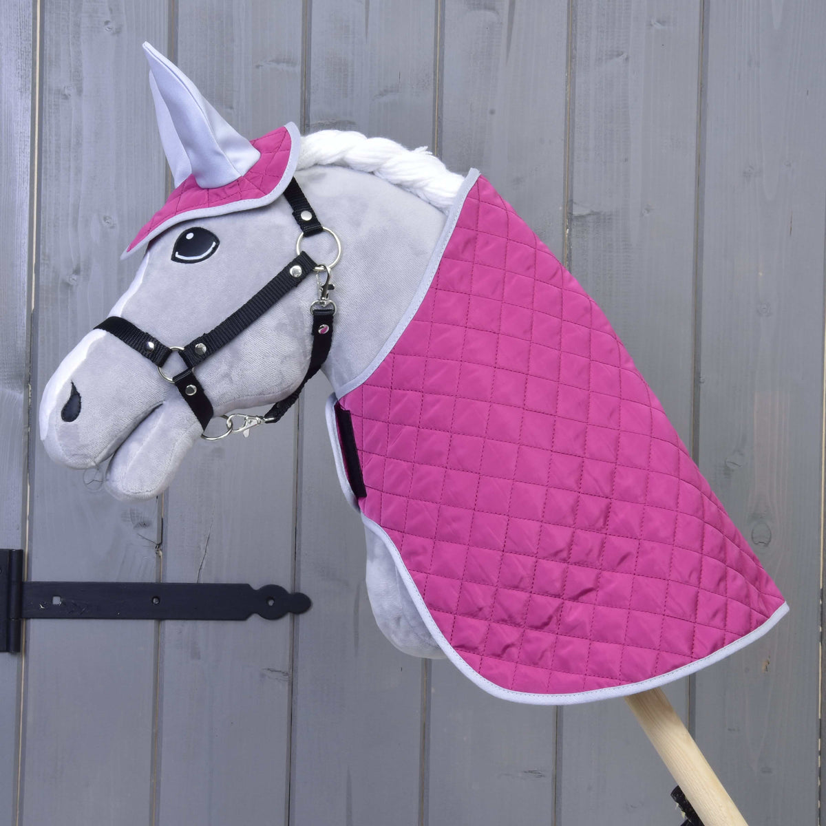 The Hobby Horse LarDen Set Hot Pink Silver features a plush hobby horse with a gray face, black bridle, white mane, quilted hot pink and white blanket set, and matching ear bonnet, all mounted on a wooden stick against a wooden wall.