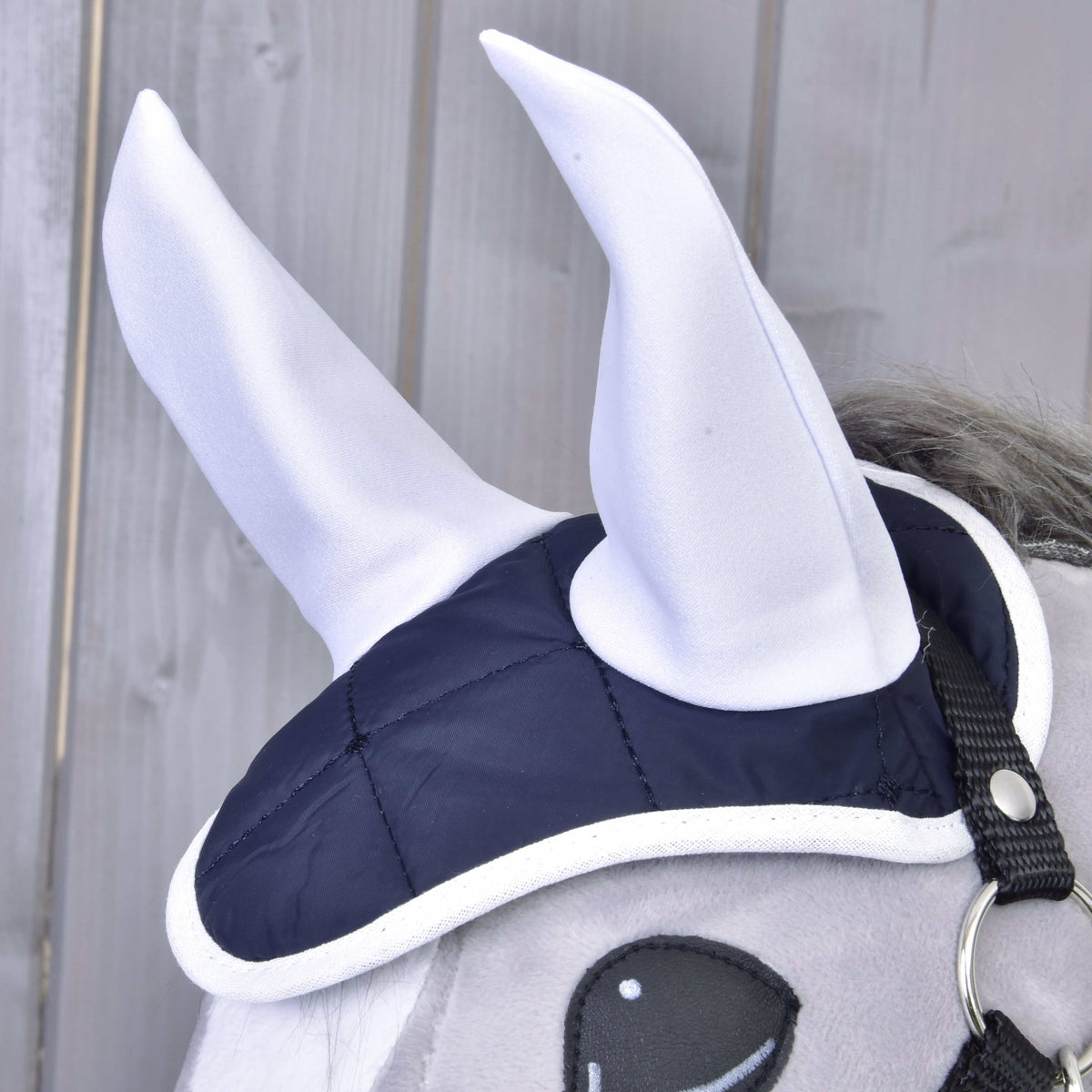 Set Monte by Hobby Horse LarDen: A gray plush horse head features a navy blue and white ear bonnet set with two large, white, pointed fabric horns, shown against wooden paneling for a striking look.