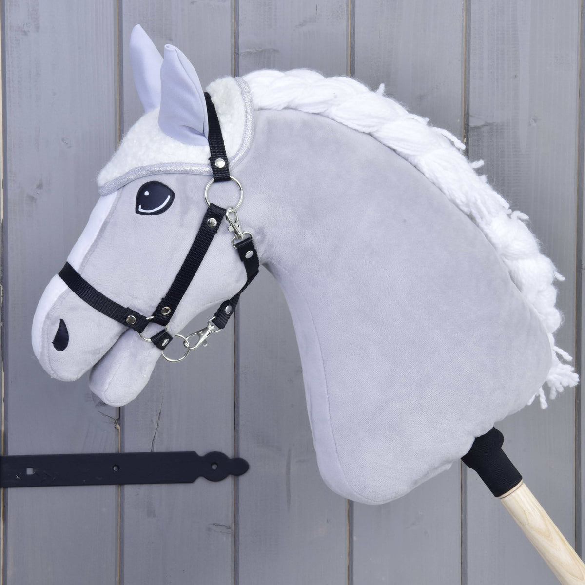 A close-up of the Hobby Horse LarDen Set Snowflake, featuring a gray body, white mane, black bridle and reins, with an ear bonnet on a wooden stick, set against a gray wooden background.
