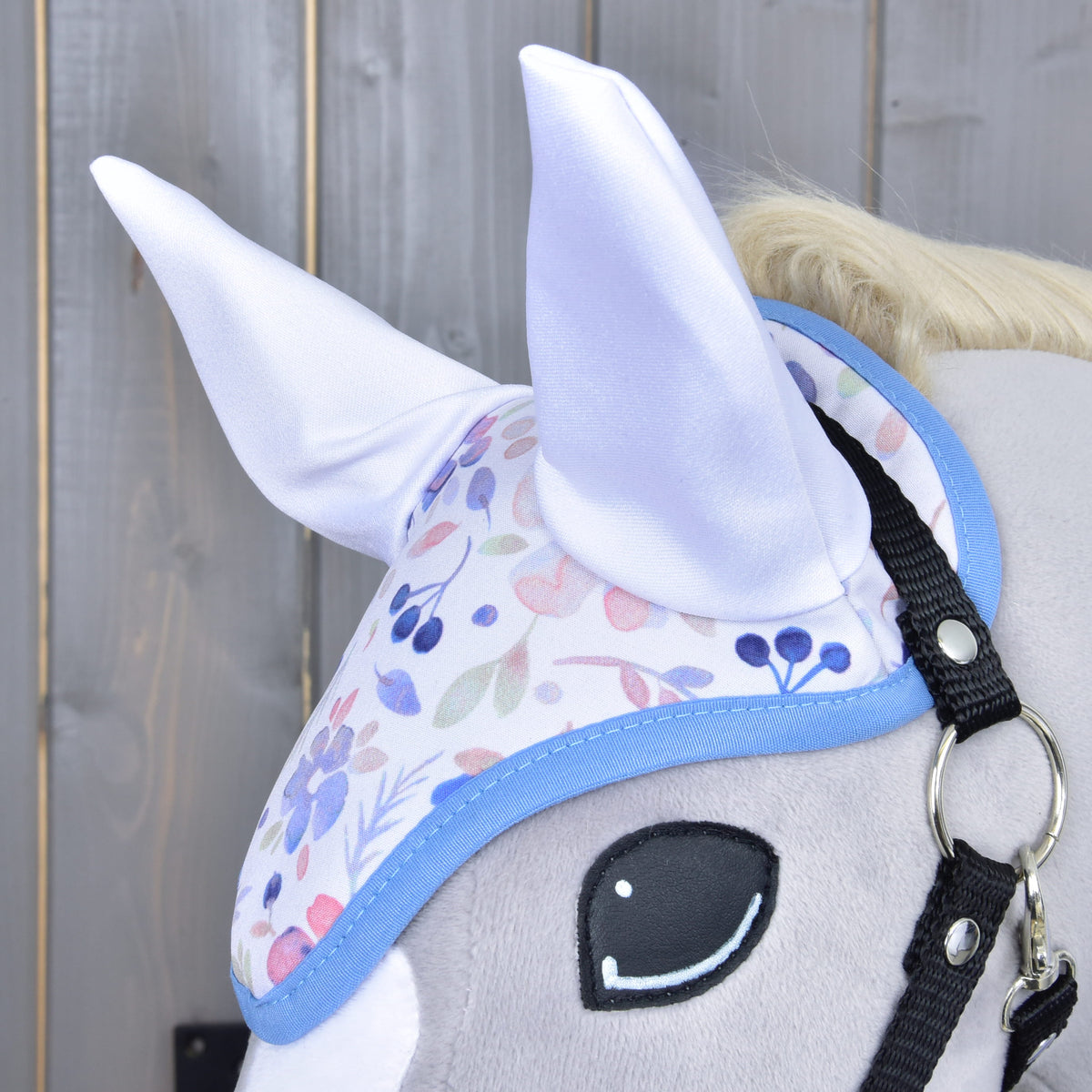 Close-up of the Set Vanessa NEW by Hobby Horse LarDen: a plush hobby horse head with a pastel floral ear bonnet featuring soft white ears, shown against a wooden background.