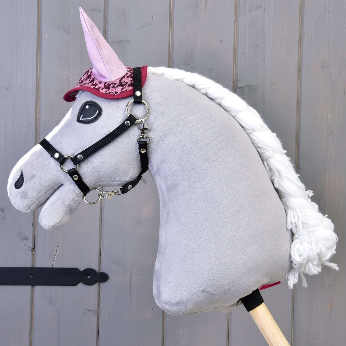 The Hobby Horse LarDen Set festive Bordeaux features a plush grey hobby horse with white mane, pink ears, and red bridle, wearing a stylish blanket, mounted on a wooden stick against a gray wooden background.