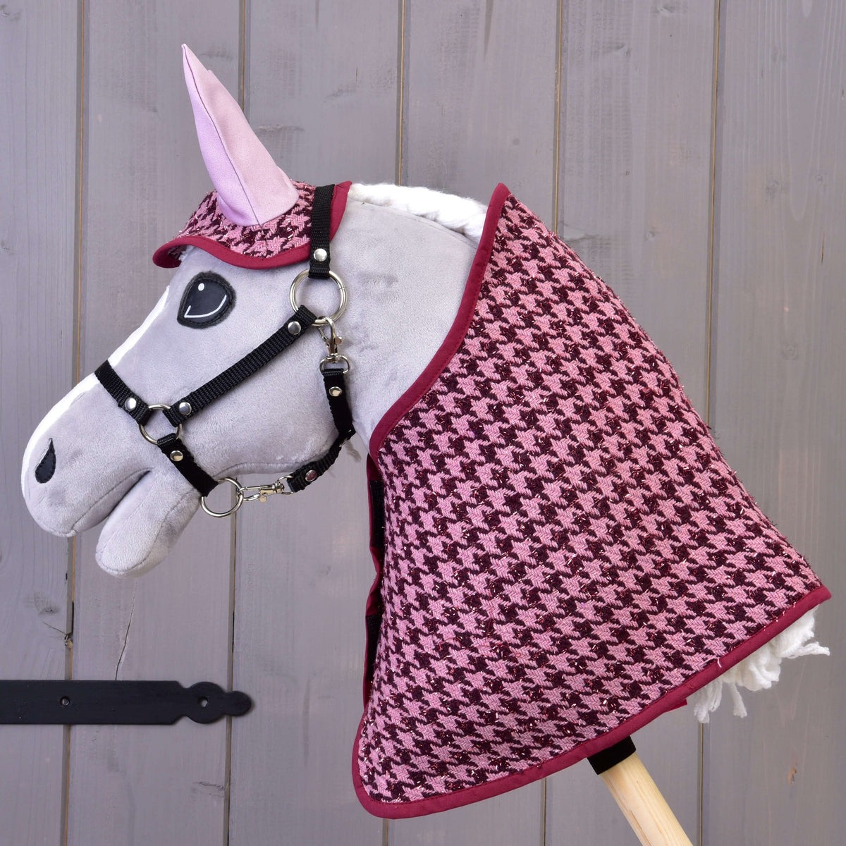 A gray stick horse toy by Hobby Horse LarDen wears the Set festive Bordeaux pink and black houndstooth blanket and ear bonnet set, perfect as a horse decoration, posed against a gray wooden wall.