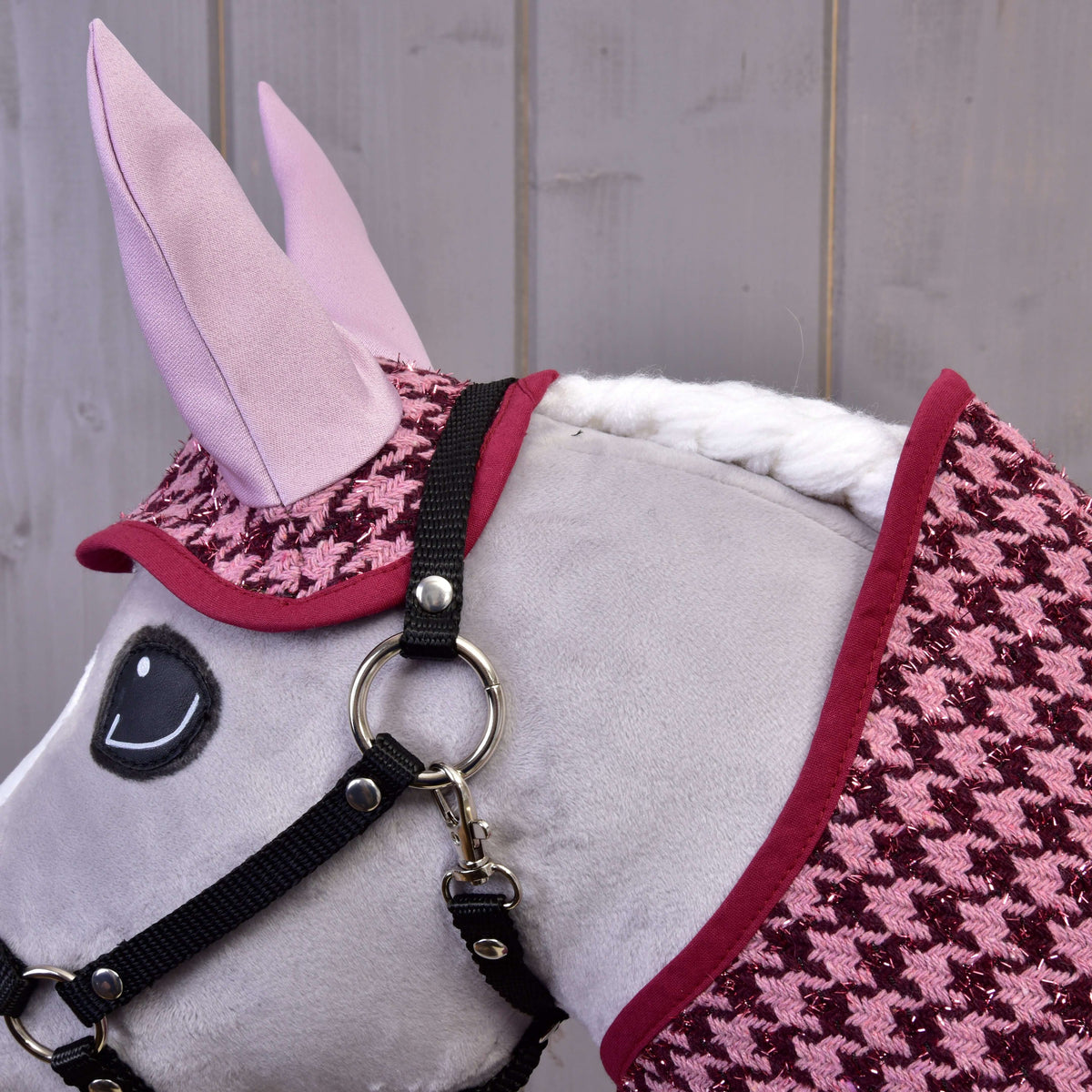 A close-up of a plush toy horse wearing a black bridle and Hobby Horse LarDen’s Set festive Bordeaux blanket and ear bonnet, set against a wooden wall, creates a charming horse decoration.