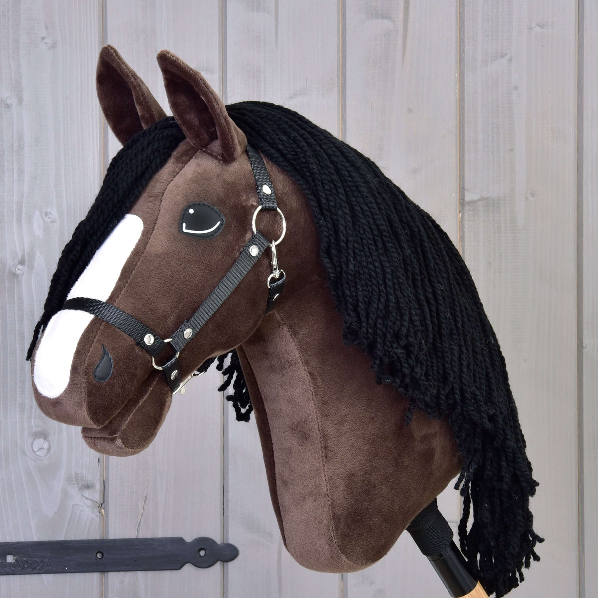 The Hobby Horse LarDen Hobby Horse Sisi with Black Halter, featuring dark brown fur, a white facial stripe, black yarn mane, and resembling a young Hanoverian sport horse, is displayed against a wooden panel background.